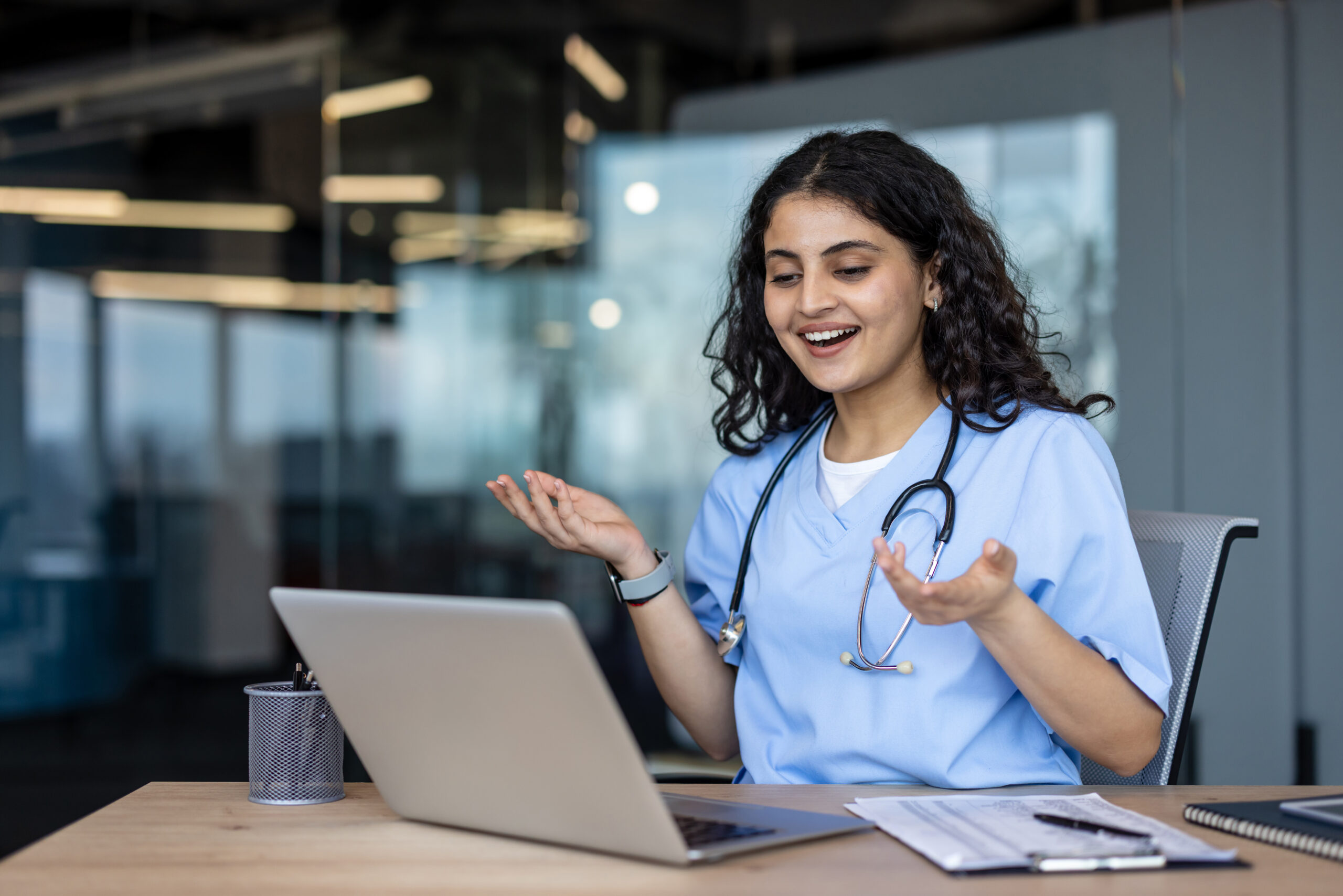 Young indian female doctor in scrubs and stethoscope smiling and gesturing while having a video call on her laptop, providing an online medical consultation from a modern office
